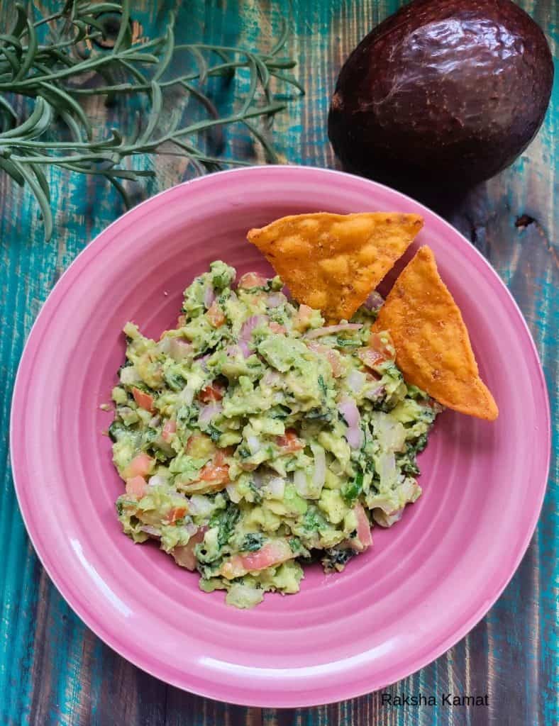 Fresh homemade guacamole served in a pink bowl with tortilla chips, featuring diced avocado, tomato, onion, and cilantro, with a whole avocado and herbs in the background.