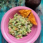 Chunky guacamole made with avocado, tomato, onion, and herbs, served in a pink plate with crispy nacho chips, placed on a rustic teal wooden background.