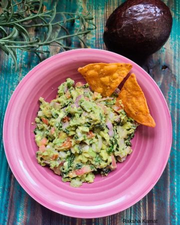Chunky guacamole made with avocado, tomato, onion, and herbs, served in a pink plate with crispy nacho chips, placed on a rustic teal wooden background.