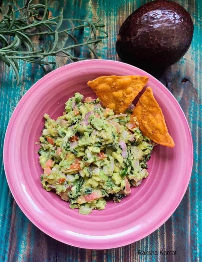 Chunky guacamole made with avocado, tomato, onion, and herbs, served in a pink plate with crispy nacho chips, placed on a rustic teal wooden background.